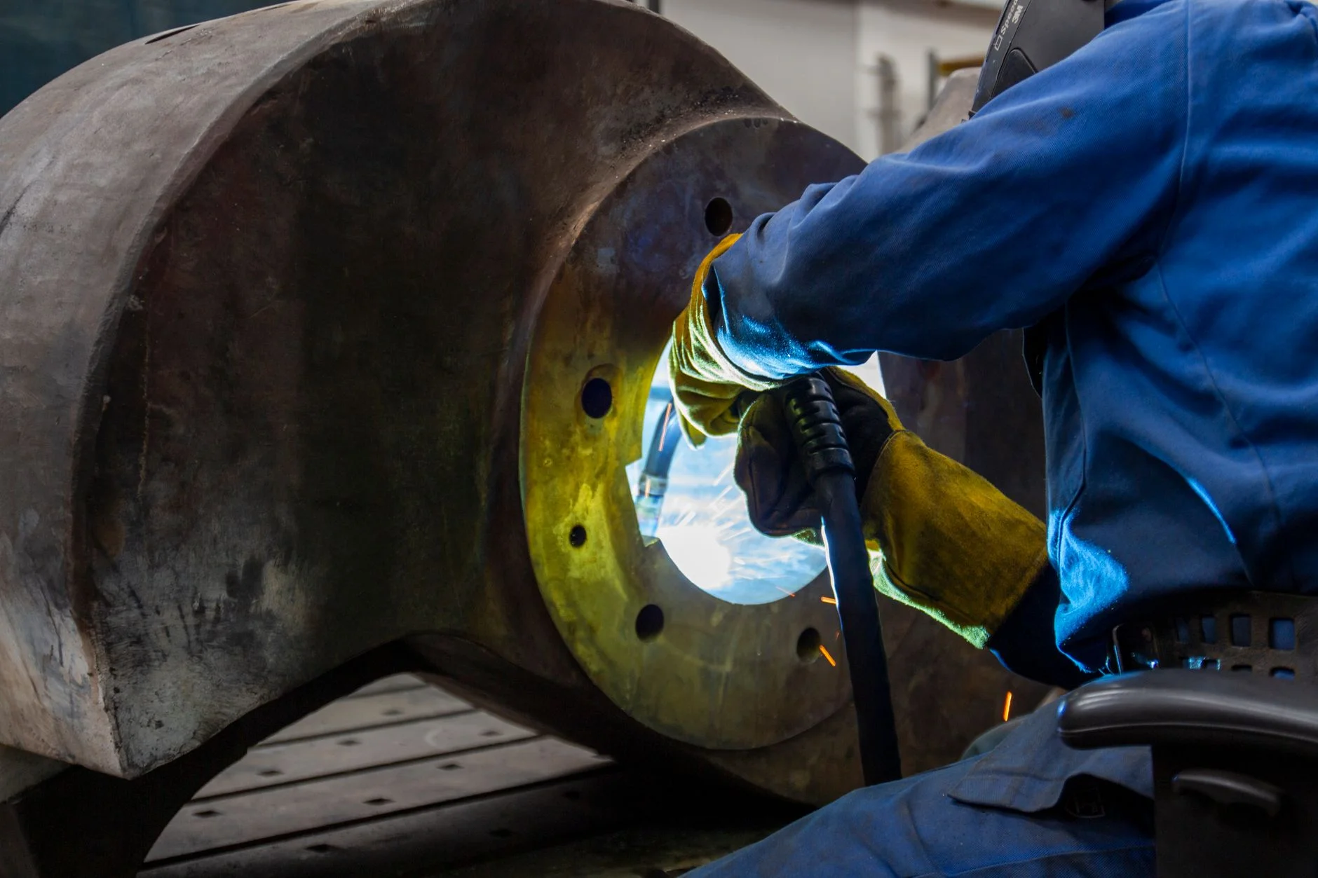 A welder working inside a large flanged cylindrical component in a fabrication shop, sparks visible against the bolt-hole face