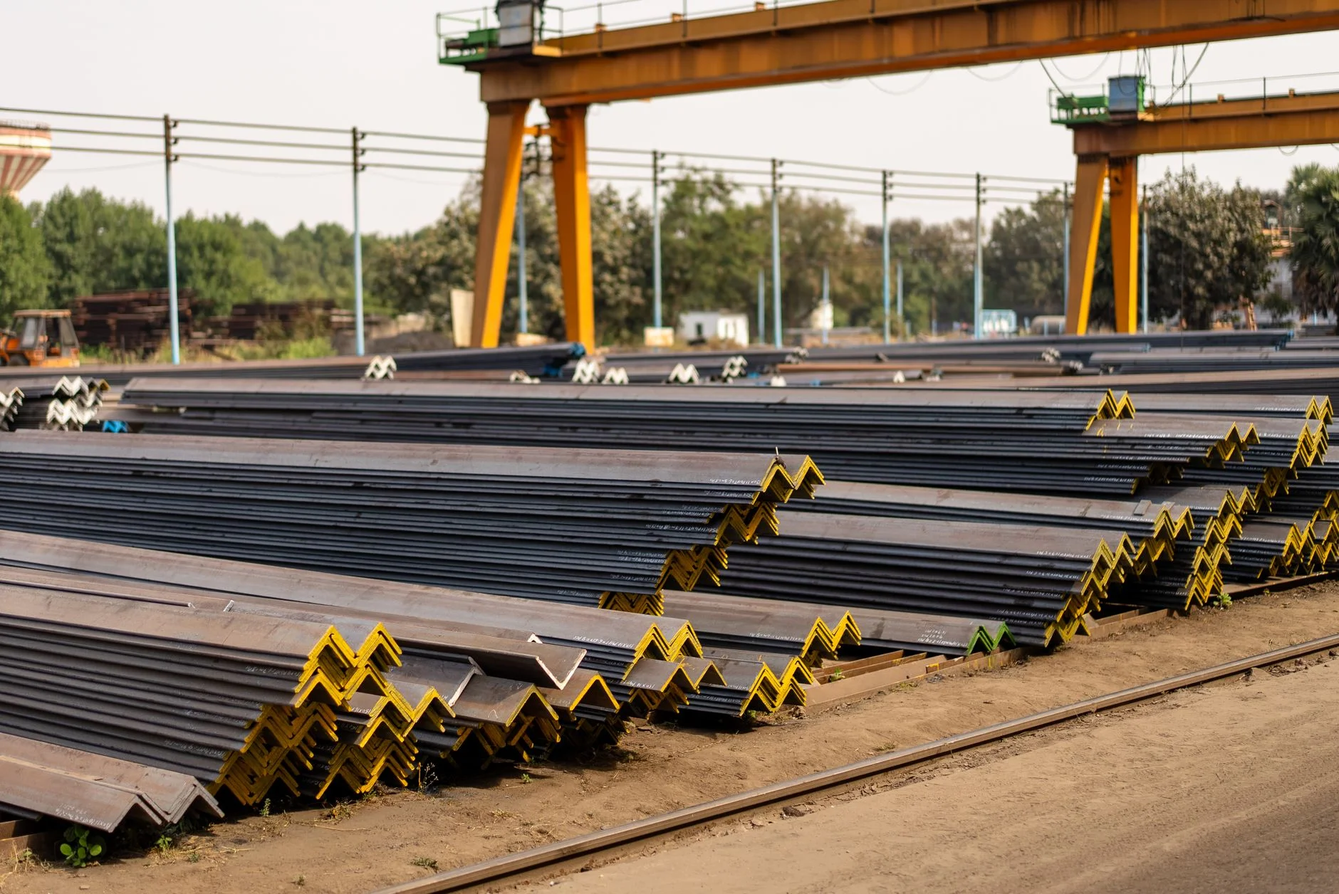 Stacked steel angle sections in an outdoor stockyard under a gantry crane, raw material waiting to become line items