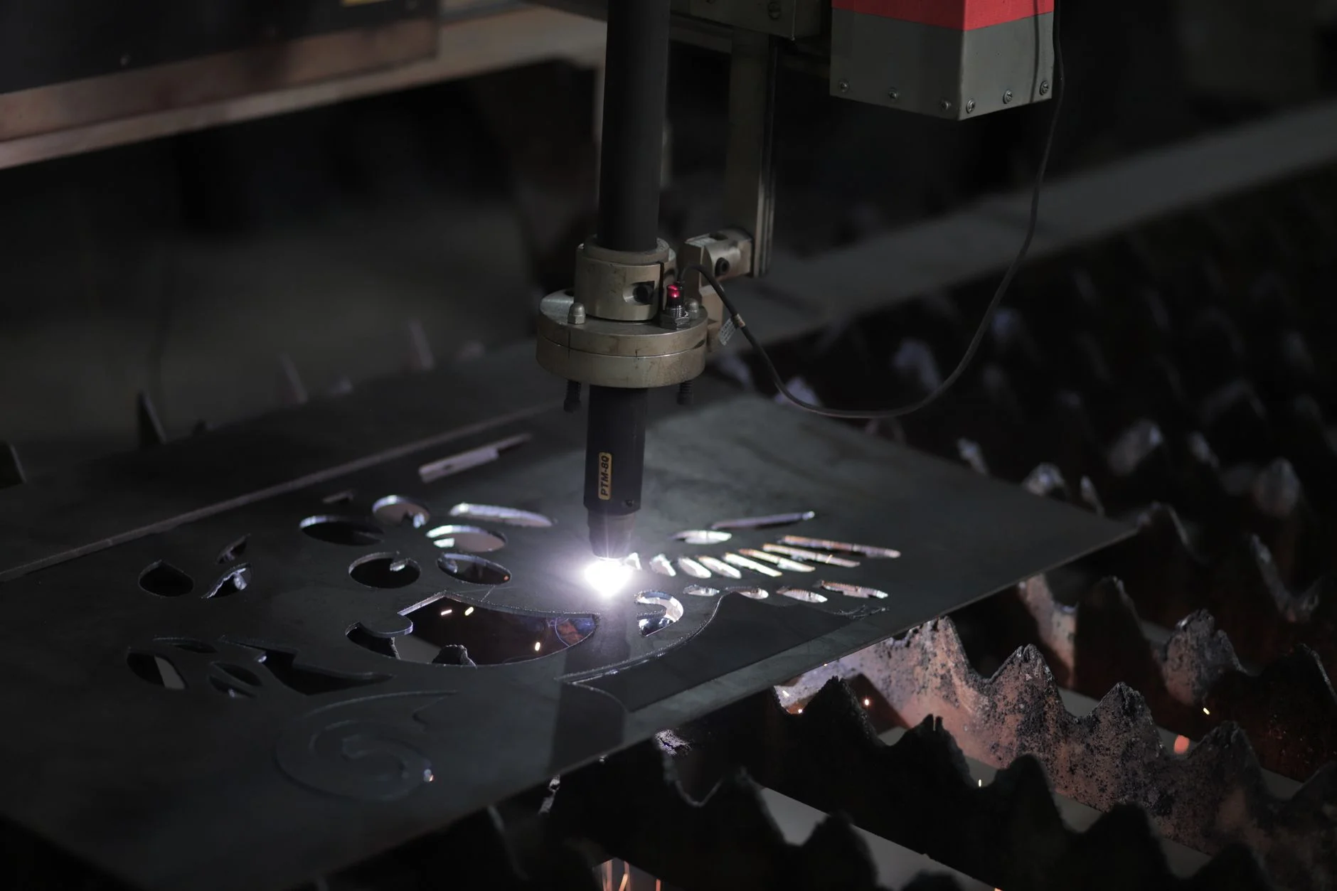 A CNC plasma head cutting profiles from a steel sheet on a slat table, cut shapes already dropped out around it